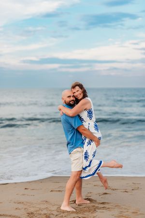 A couple embraces joyfully on a sandy beach, the woman in a blue floral dress lifted by a man in a blue shirt.