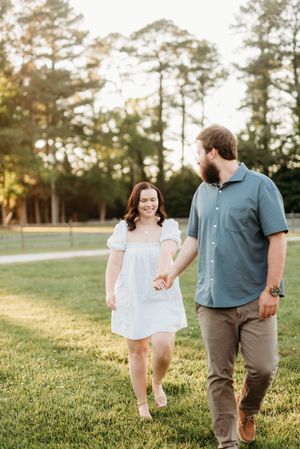 A couple walks hand-in-hand through a sunlit park, woman in white dress, man in blue shirt and khaki pants.
