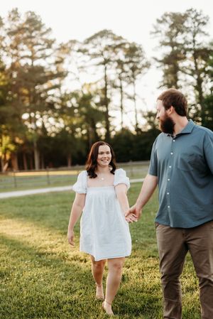 Couple holding hands walking through green field at golden hour, woman in white dress smiling.