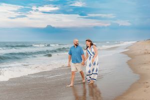 Couple walking hand-in-hand on a sandy beach with ocean waves, man in blue shirt and woman in floral dress.