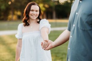 Smiling woman in white dress holds hands with partner outdoors in a sunny park during an engagement photo session.