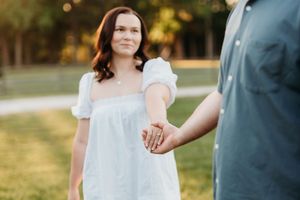 A woman in a white dress holds hands with a man outdoors in a sunny park setting.