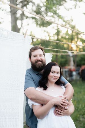 Bearded man embracing woman in white dress outdoors near hanging white lace fabric at sunset.