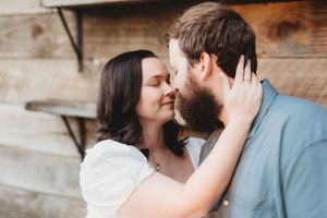 A couple shares a romantic almost-kiss moment, woman in white top holding mans bearded face, against rustic wood wall.