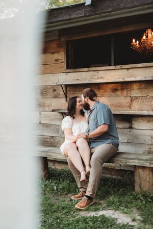 A couple shares a romantic moment sitting on a rustic wooden bench outside a log cabin with a chandelier visible inside.