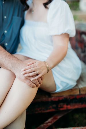 Couple holding hands tenderly, woman in white dress with gold bracelet, man in blue shirt, seated on rustic bench.