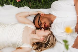 Couple lying on white blanket in grass, sharing a romantic moment, woman in white dress, man in white shirt.