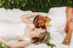 Couple in white attire lying on a blanket in lush green grass, sharing a romantic moment outdoors.