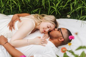 A romantic interracial couple lying on a white blanket in green grass, sharing a tender moment with flowers nearby.