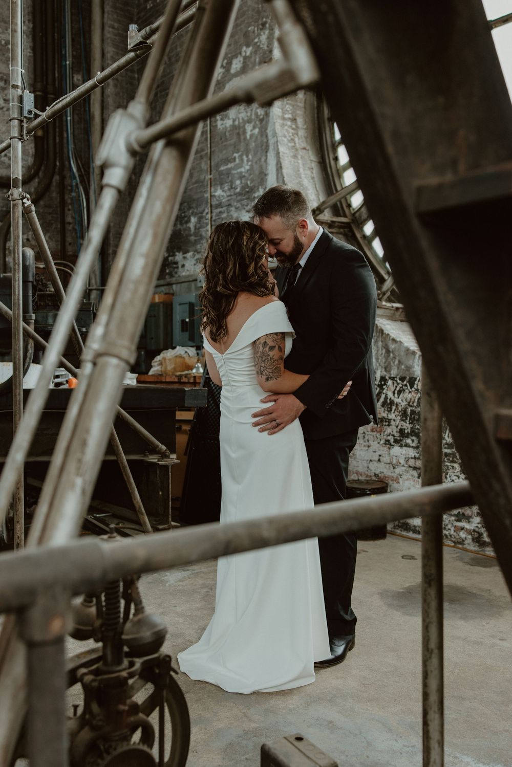 A bride and groom share a romantic kiss in an industrial wedding venue, bride wearing an off-shoulder gown with a tattoo.