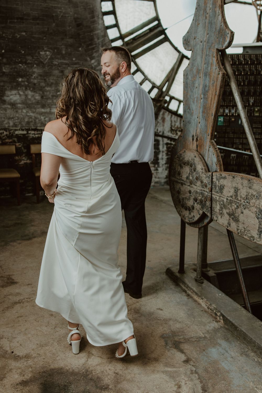 Bride in white off-shoulder gown and groom in black trousers share a joyful moment near a vintage clock tower.