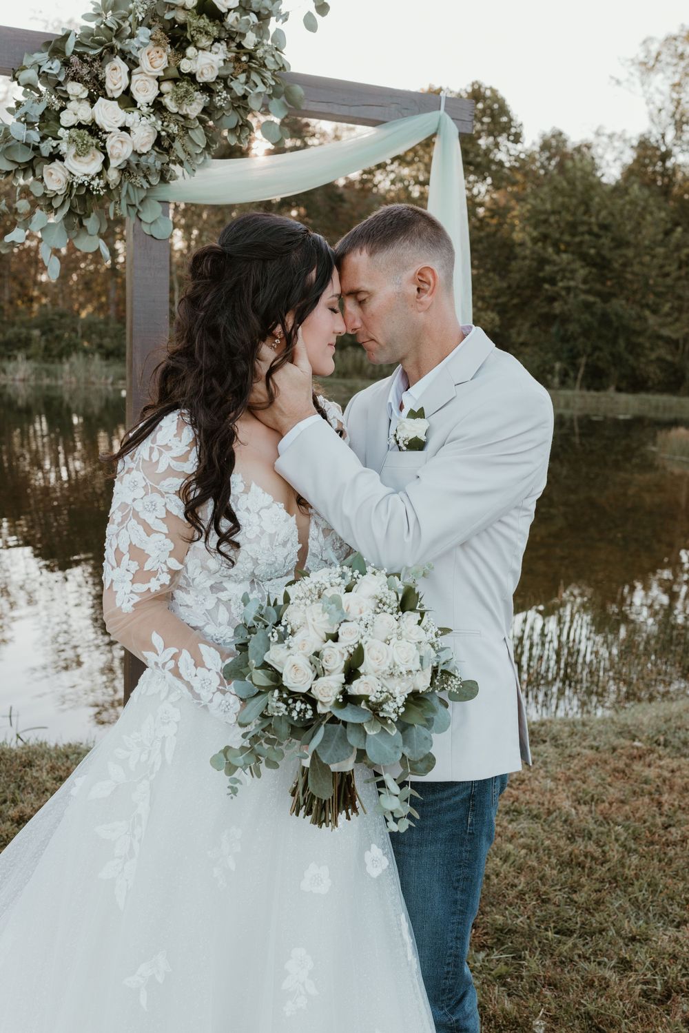 Bride and groom share romantic moment under floral arch by pond.