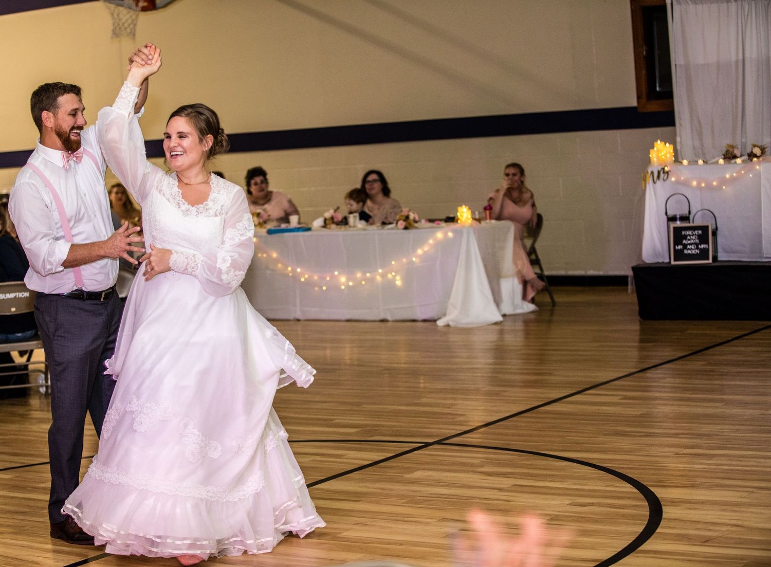 A couple dances on a wooden gymnasium floor during a wedding reception with tables and guests visible in the background.