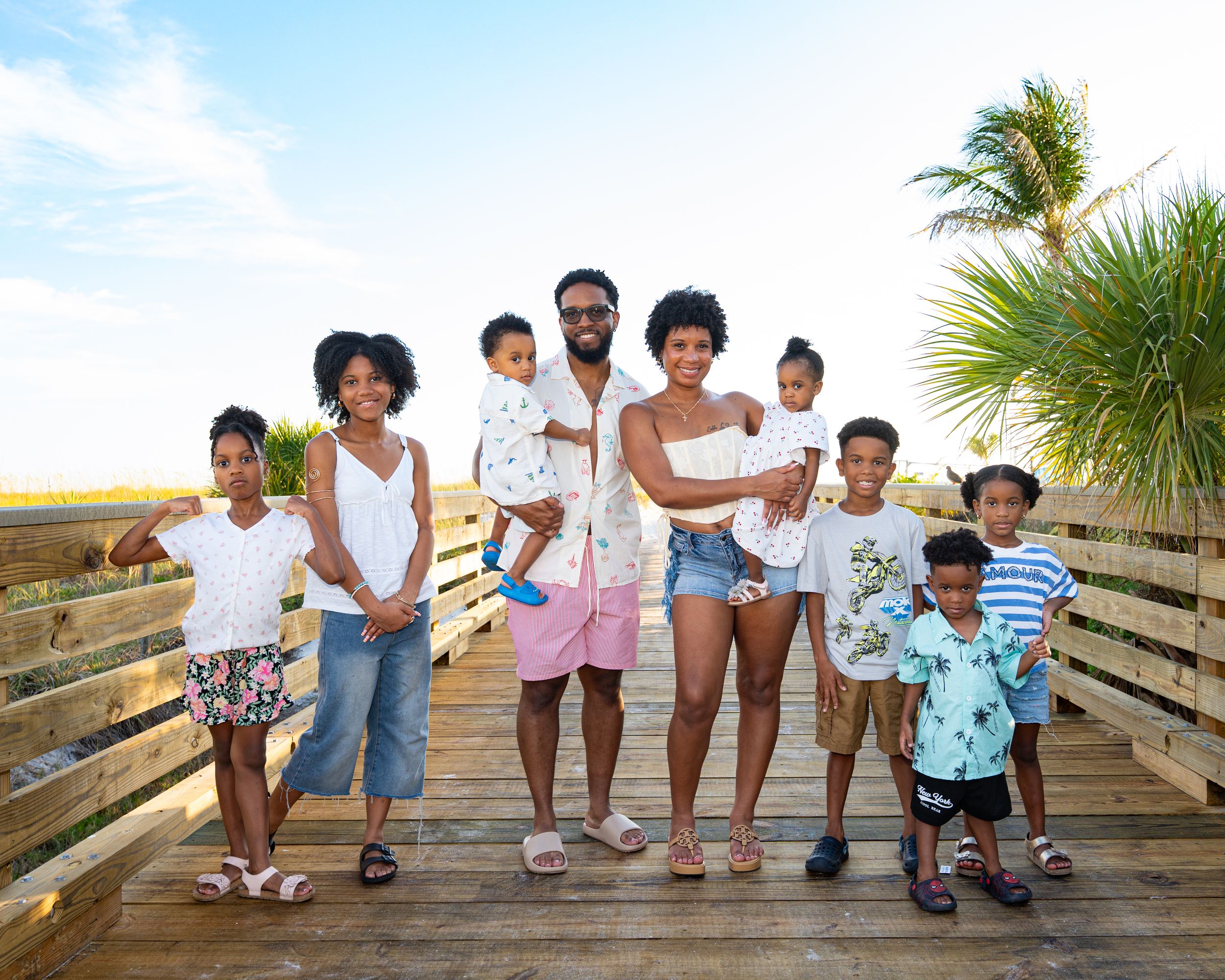 Happy Black family of seven posing together on a sunny beach boardwalk with palm trees in the background. St Pete Beach-BE Jazy Photography