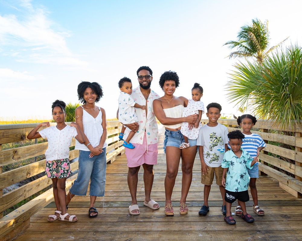 Happy Black family of seven posing together on a wooden boardwalk near a beach on a sunny day.