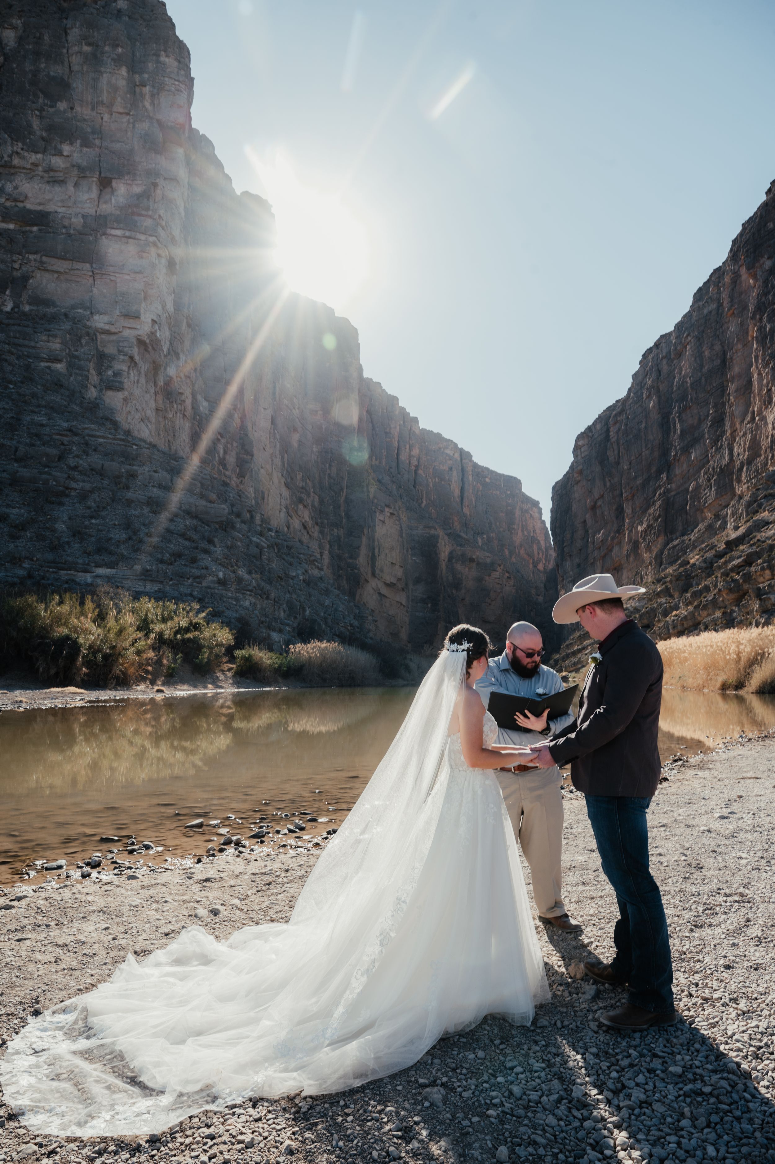 A wedding ceremony takes place along a riverbank in a dramatic canyon with towering rock walls and bright sunlight.