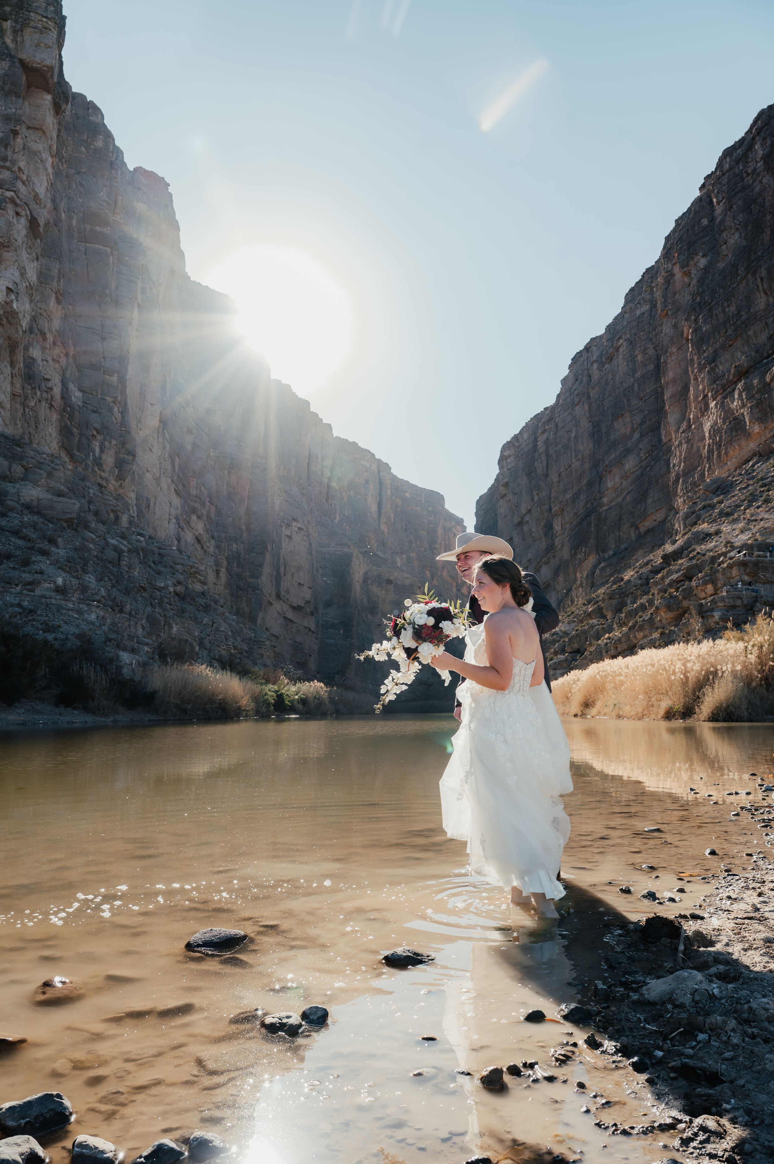 A bride in a flowing white dress stands at the edge of a river canyon with dramatic cliffs and sunlight peeking through.