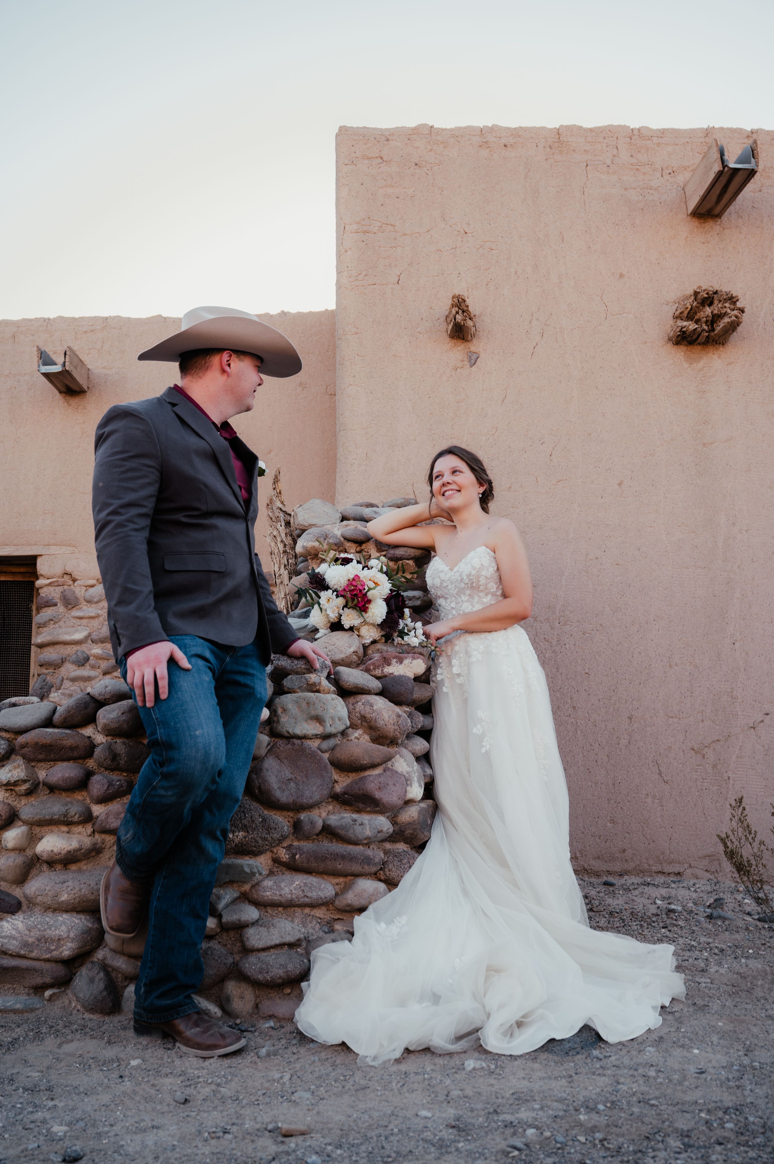 A couple in western wedding attire pose against a rustic adobe wall with stone base during a Southwestern ceremony.