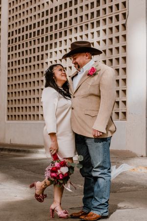 A romantic wedding moment showing a couple in white and tan embracing against a textured wall with red flowers.