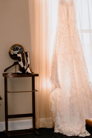 A delicate lace wedding dress hangs by a window next to vintage decor.