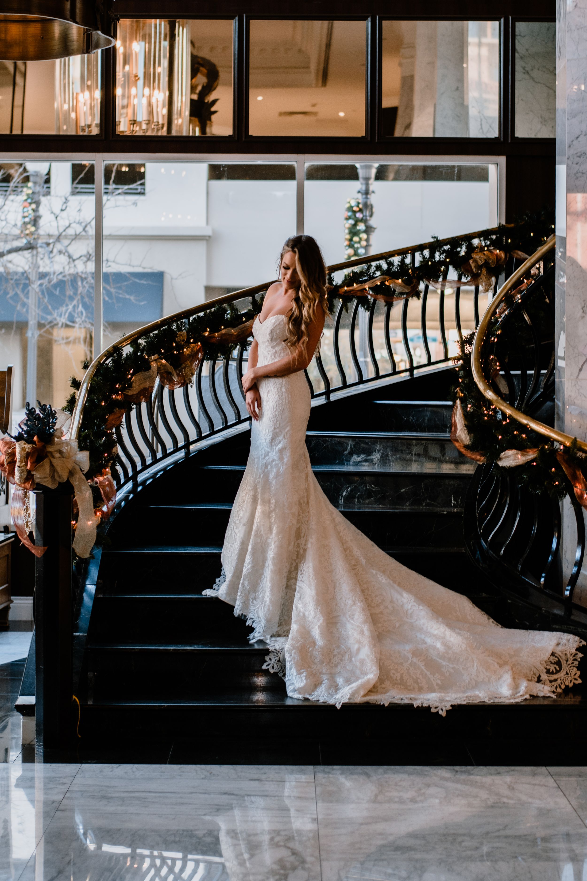 A bride in a lace wedding gown poses on an elegant curved staircase decorated with lights.