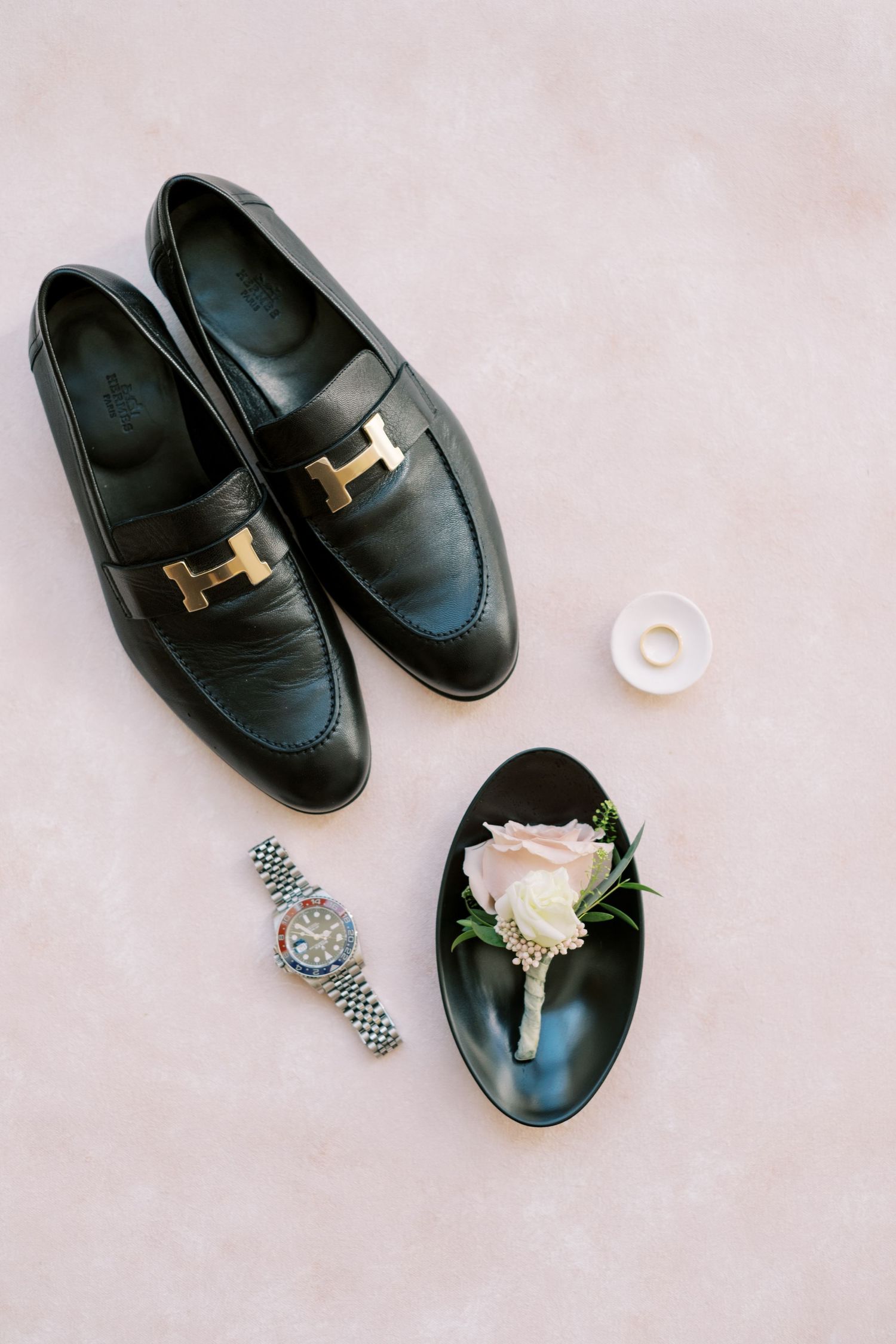 Black leather dress shoes with accessories and a rose on light pink background.