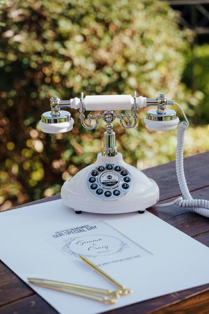 Vintage white rotary telephone with guest book and pencil on wooden table in outdoor garden setting.