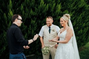 A wedding party gathers outdoors for candid moments against a backdrop of lush green hedges.