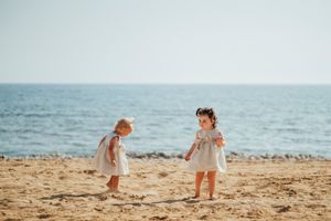 Two children in light summer dresses play together on a sandy beach with the ocean in the background.
