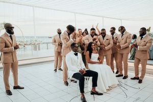 Wedding party in matching tan suits gathers on a rooftop terrace overlooking a waterfront venue during celebration.