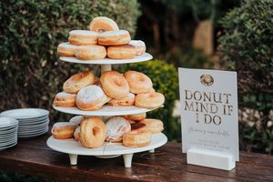 A tiered display of glazed donuts on a white stand next to a 'Donut Mind If I Do' sign at an outdoor event.