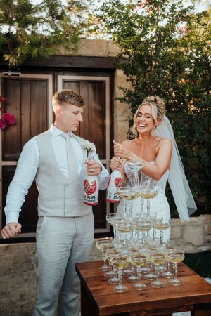 A couple pours champagne into a tower of glasses during their outdoor wedding celebration.