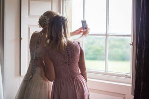 Two people look out a sunny window in elegant dresses during wedding preparations.