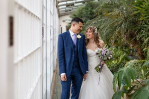 Couple in wedding attire pose together along a white wall with tropical plants.