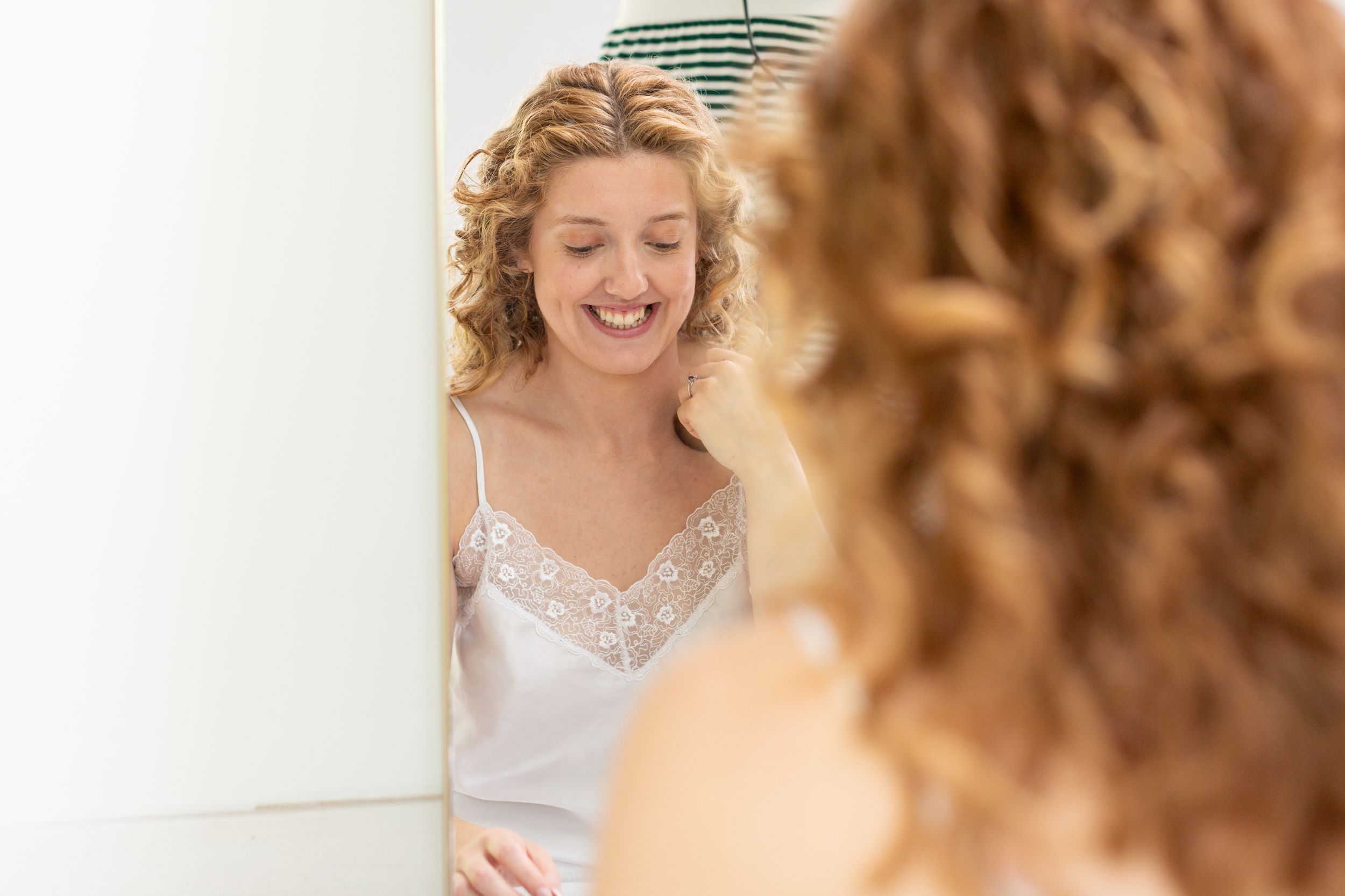 A reflection in the mirror shows someone wearing a white lace-trimmed camisole while smiling and looking down.