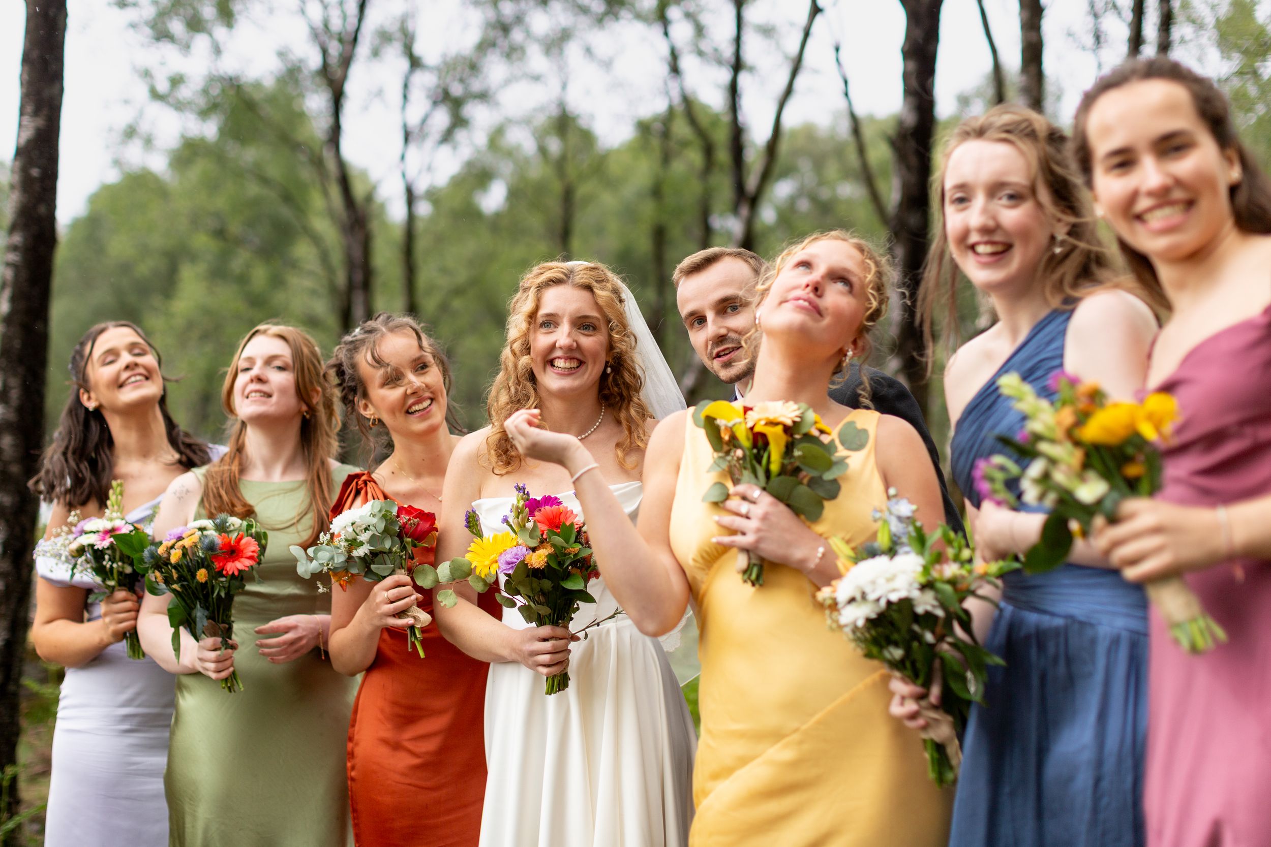 Wedding party in colorful dresses laughs together while holding bouquets in an outdoor garden setting.