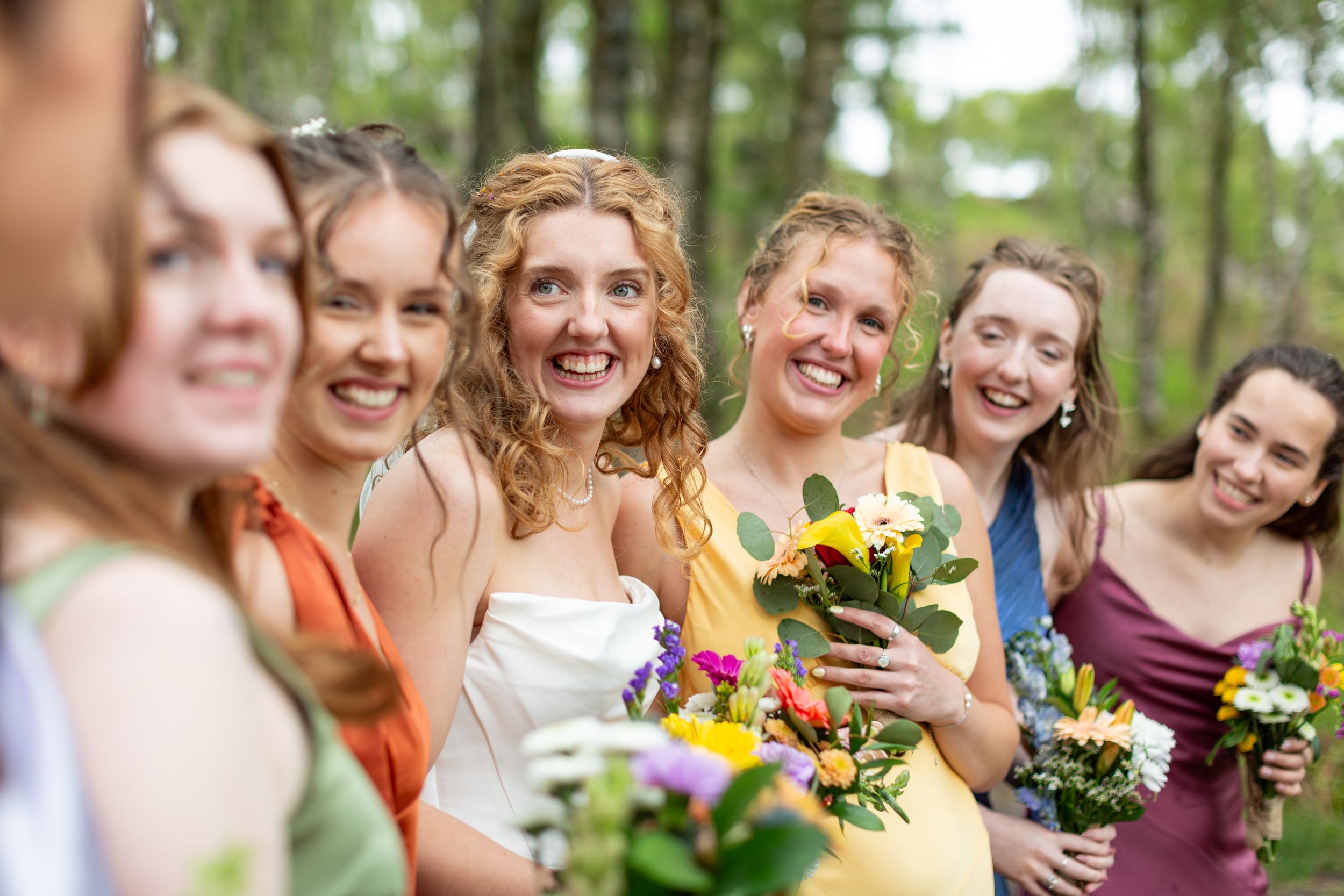 A group of bridesmaids in burgundy and yellow dresses laugh together while holding colorful bouquets outdoors.