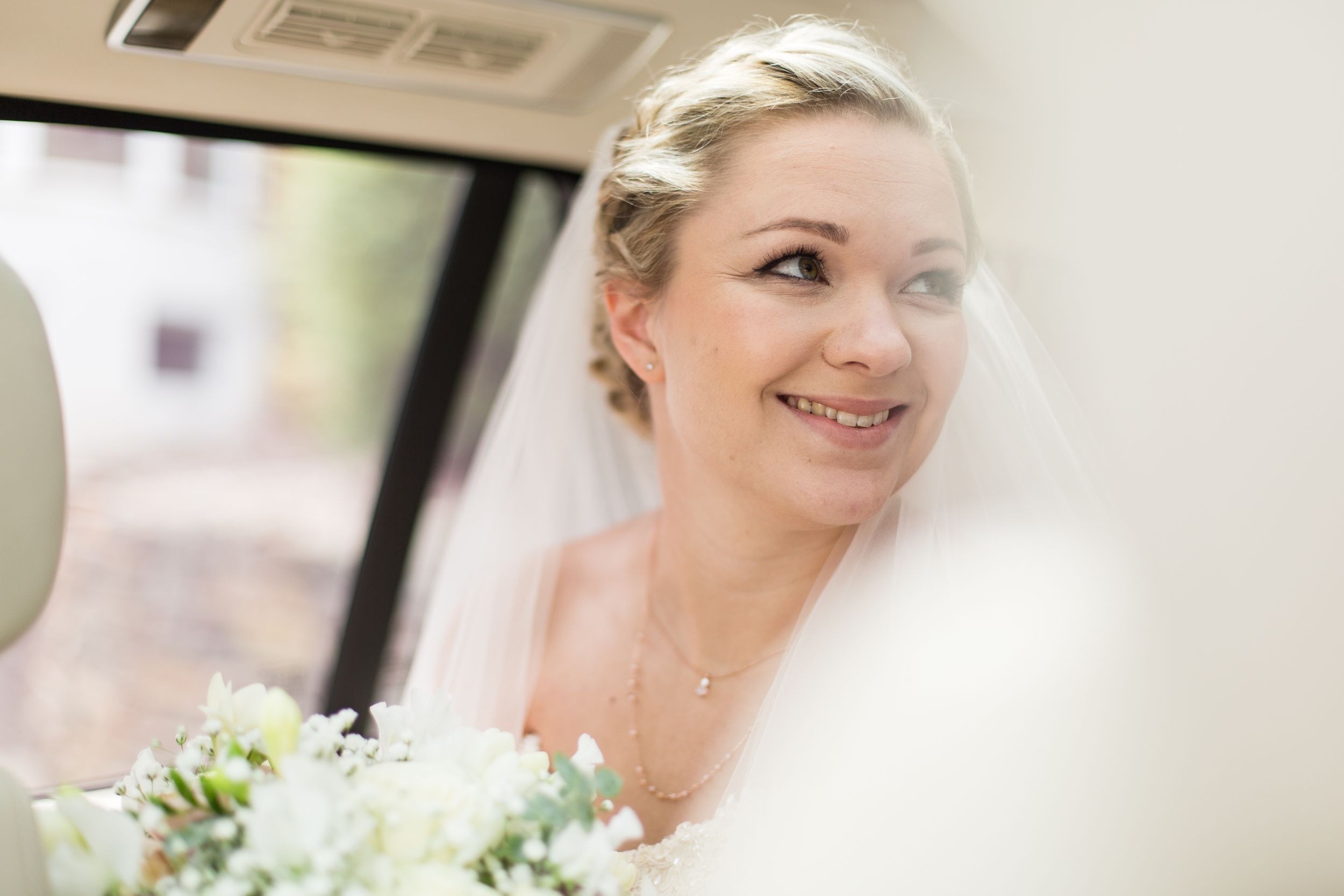 A bride wearing a traditional white veil smiles while holding a bouquet of white flowers on her wedding day.