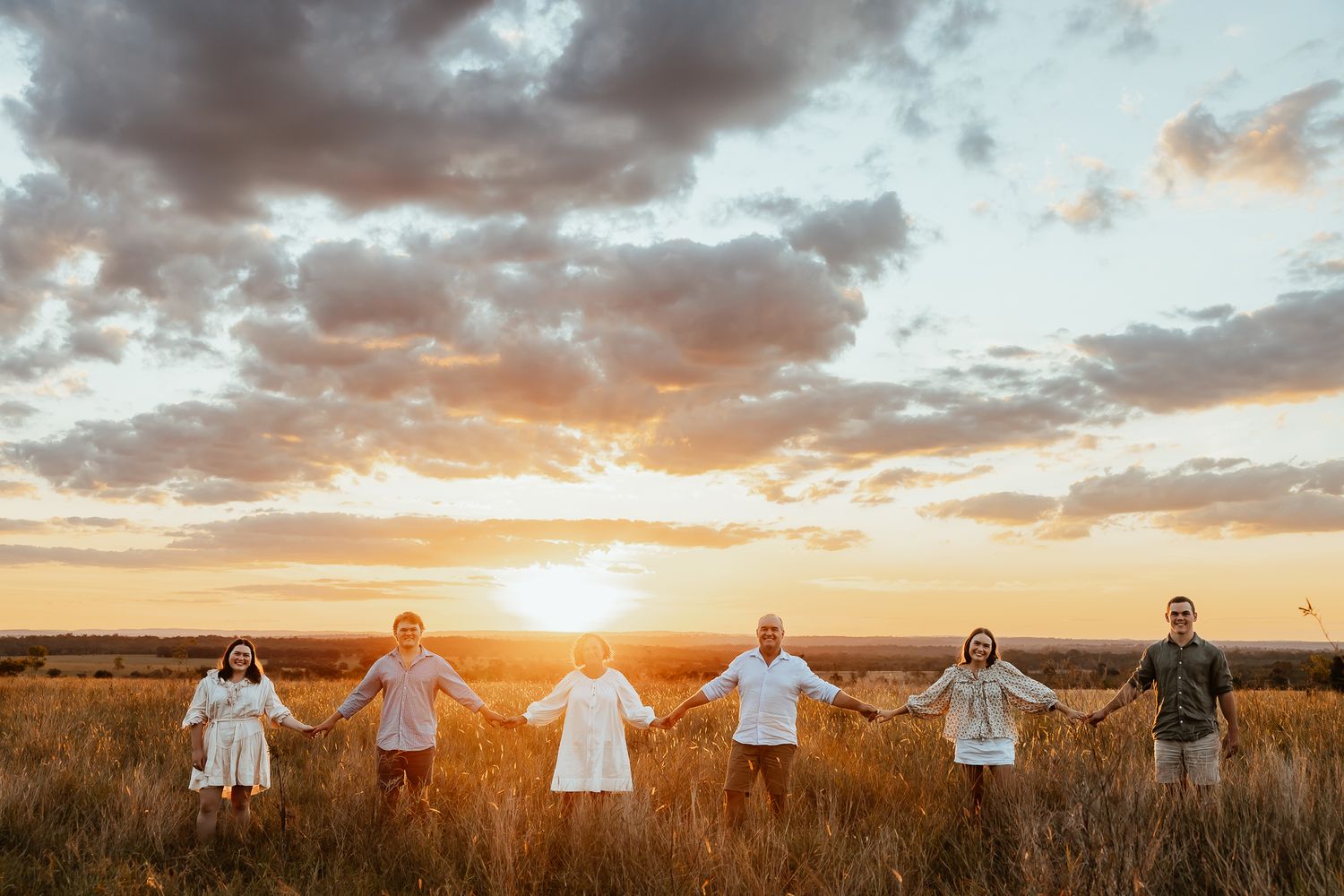 Six people holding hands in a line at golden sunset in an open field with dramatic clouds in the background.