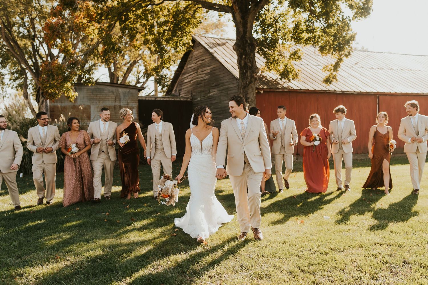 A couple holding hands and smiling at each other. They are standing with their wedding party, which is blurry in the background.