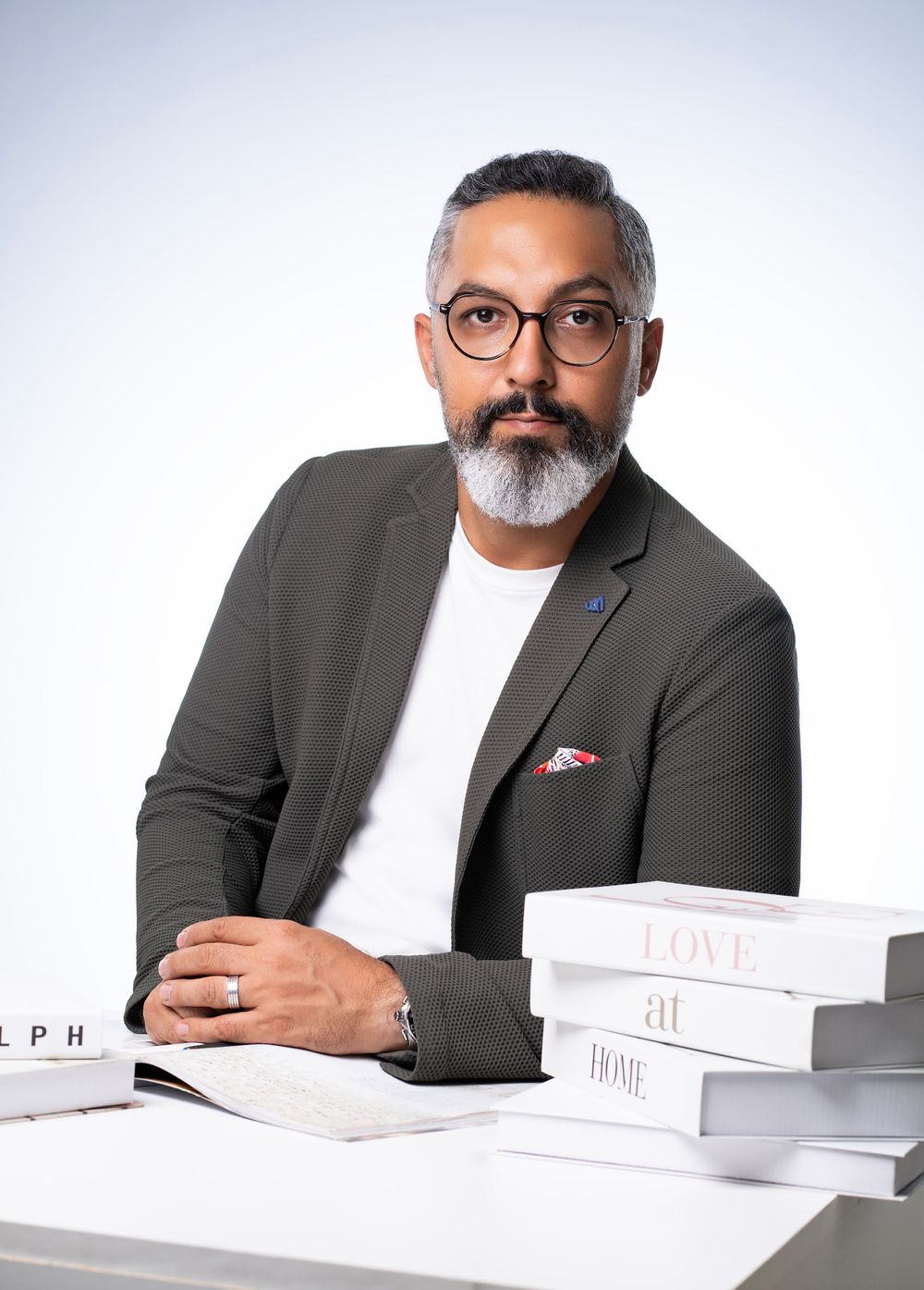 Professional with glasses and gray beard wearing blazer at desk with stacked books.