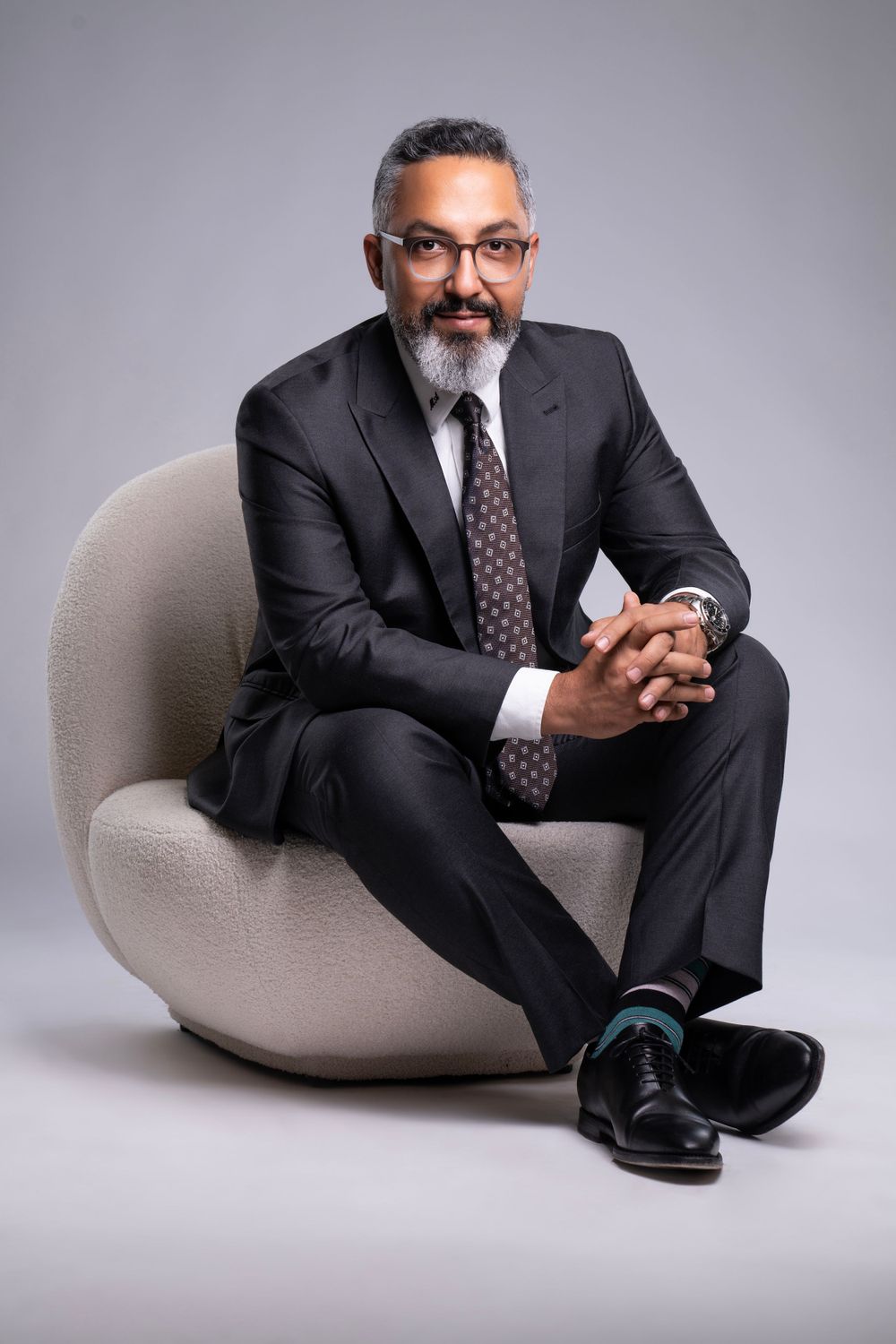 Professional in black suit seated in modern gray armchair against studio background.