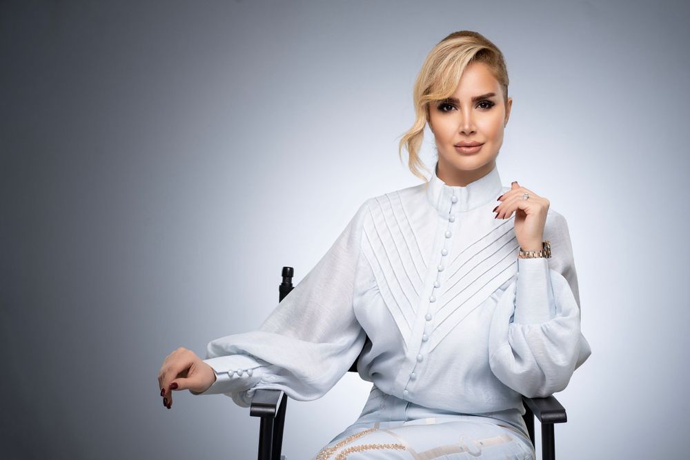 Professional woman in white ruffled blouse seated against gray background in studio setting.