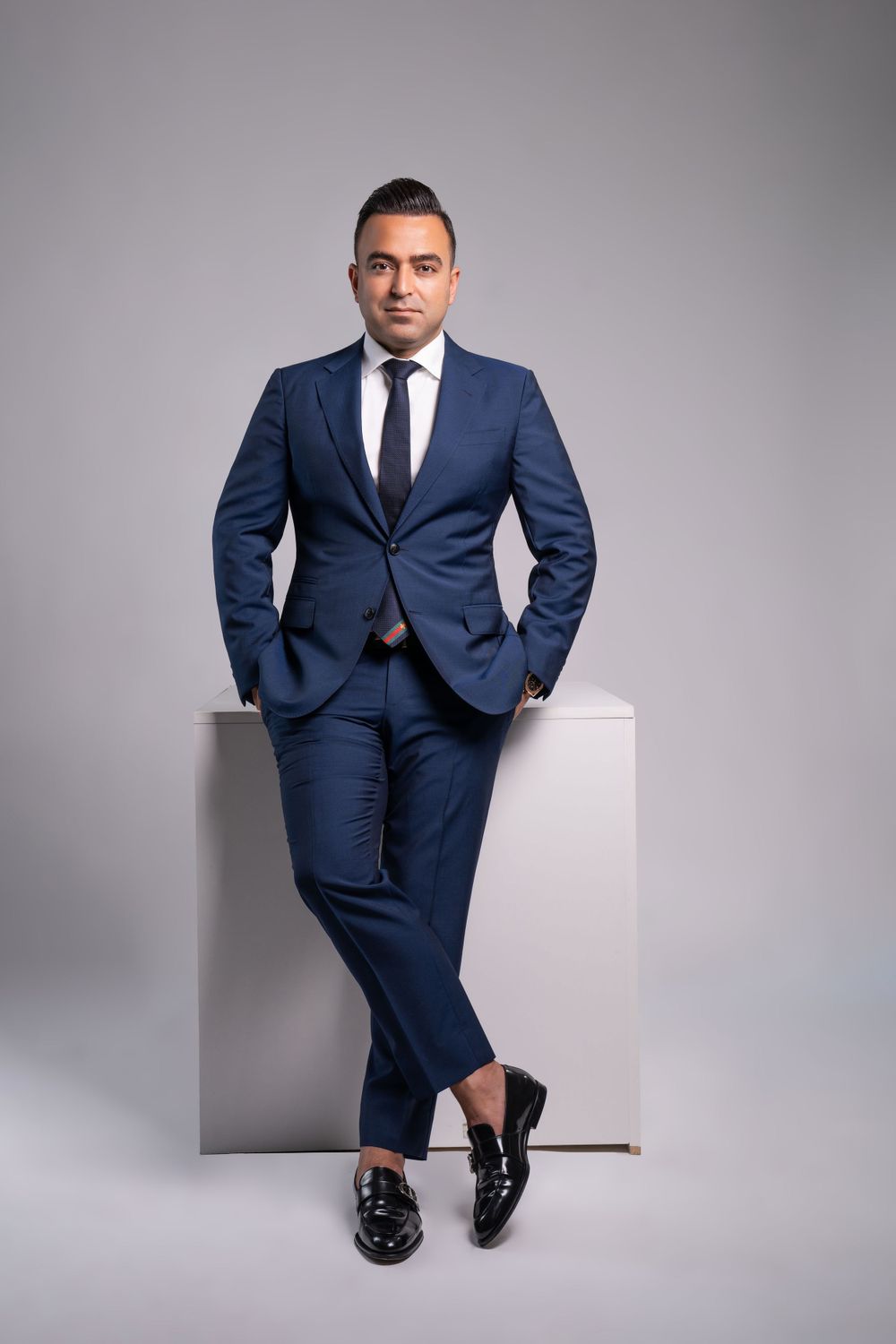 Professional in navy suit seated on white cube against gray background.