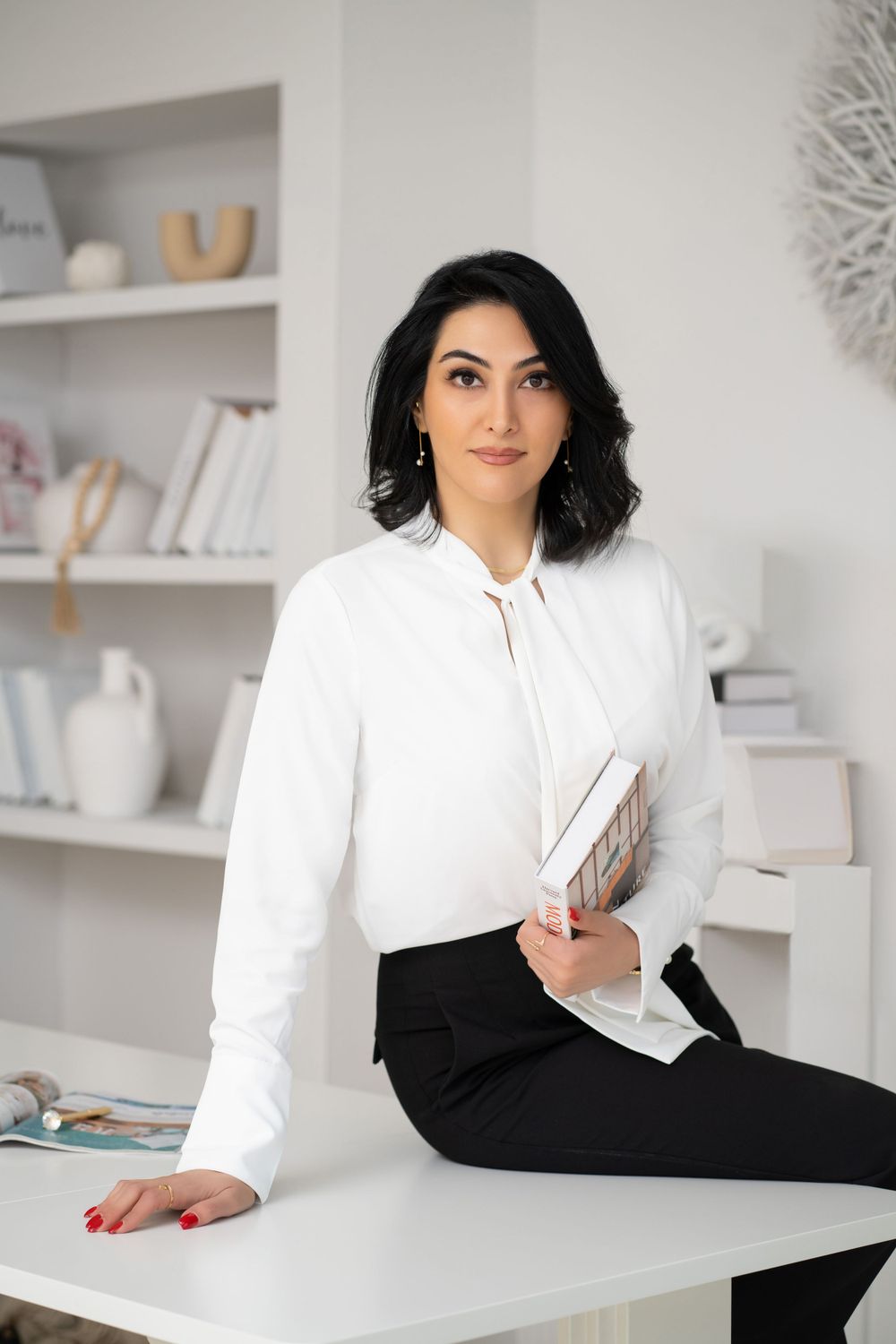 Professional in white blouse and black skirt seated on desk in modern office setting.