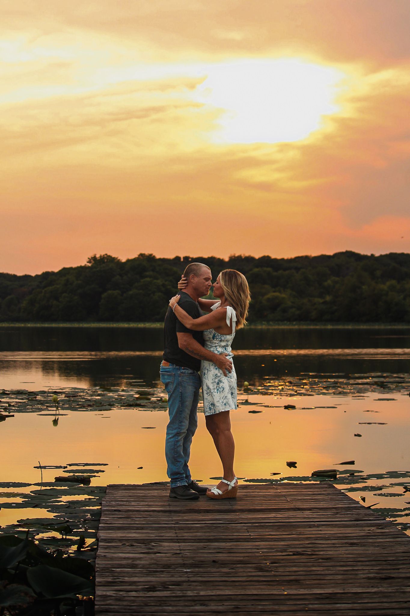Couple embracing on a wooden pier against a golden sunset over a tranquil lake.