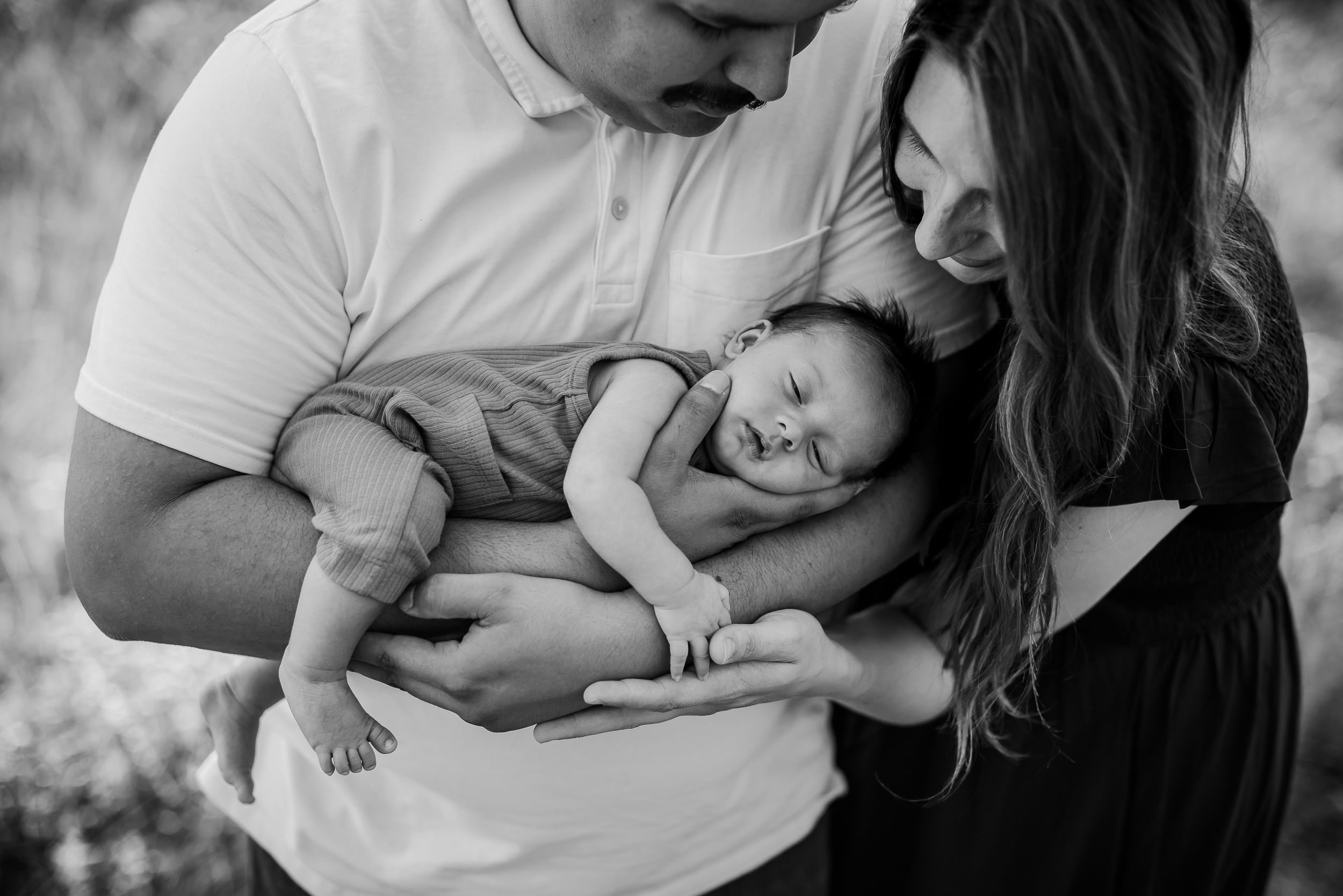 San Antonio Family Photographer. A black and white photograph showing parents tenderly holding their sleeping newborn baby outdoors..