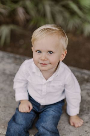 A young child in a white collared shirt and blue jeans sits outdoors in a natural setting.