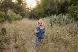 A joyful toddler girl in denim runs through a sunlit meadow path surrounded by tall grass and cacti.