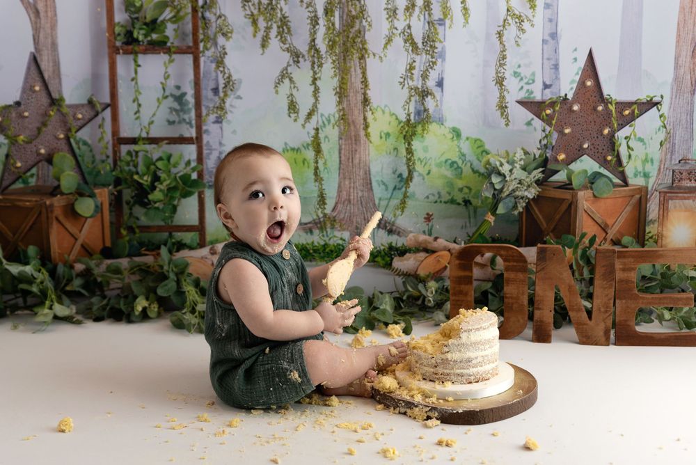 A small child wearing green sits on the floor with a birthday cake in a rustic garden-themed setting with decorative stars.